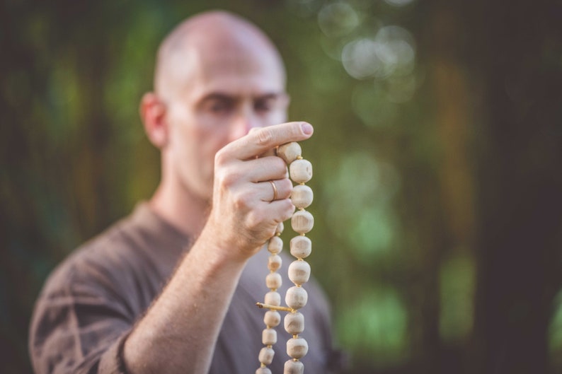 May include: A wooden mala bead necklace with large, rounded beads. The beads are strung together with a gold-colored spacer. The background is blurred, with a person holding the necklace in focus.