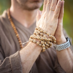 May include: A person wearing a brown shirt and a wooden bead necklace. A wrist is adorned with a light-colored watch and a bracelet of carved wooden beads. The hands are clasped together in a gesture of prayer.