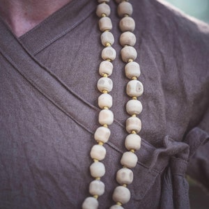 May include: Close-up of a person wearing a brown wrap-style top with a wooden bead necklace. The necklace features large, light-colored beads separated by small gold-colored spacers. The top is made of a soft, textured fabric.