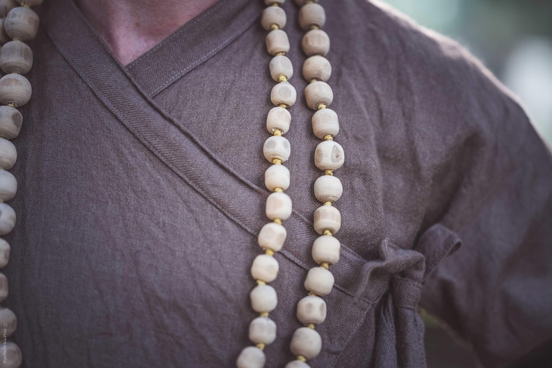 May include: Close-up of a person wearing a brown wrap-style top with a wooden bead necklace. The necklace features large, light-colored beads separated by small gold-colored spacers. The top is made of a soft, textured fabric.