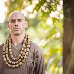 May include: A person wearing a brown wrap-style shirt and a triple-layered necklace made of cylindrical, textured light brown wooden beads with small red accents between each bead, resting against the chest in a layered, circular arrangement.