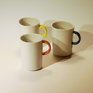 Peut inclure: Trois tasses en céramique blanche avec des anses colorées. Une tasse a une anse jaune, une autre a une anse rouge et la troisième a une anse bleue. Les tasses sont cylindriques et présentent un design simple et moderne.