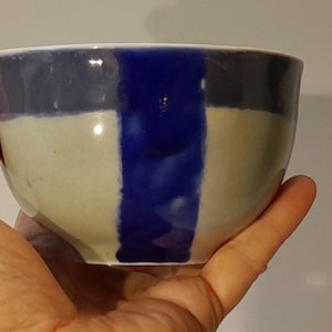 Può includere: Una ciotola in ceramica con un design dipinto a mano. La ciotola presenta una striscia blu verticale su uno sfondo crema, blu navy e bianco. La ciotola è tenuta in una mano.
