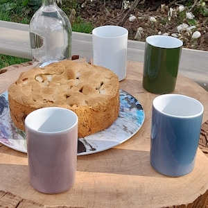 May include: Four mugs in different colours, white, green, blue and purple, are arranged on a wooden table with a plate of apple cake in the centre.