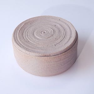 Può includere: Una scatola rotonda in ceramica di colore marrone chiaro con un coperchio. Sia il coperchio che la scatola sono decorati con un motivo a spirale concentrico.
