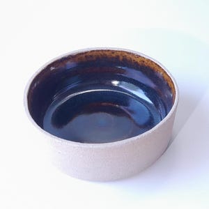 Peut inclure: Un bol en céramique fait à la main avec un extérieur beige clair et un intérieur marron foncé et bleu. Le bol a une forme simple et arrondie et une finition lisse et brillante à l'intérieur. Le bord a un aspect plus foncé et texturé.