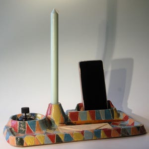 May include: A colorful ceramic desk organizer with a geometric pattern. It features a tall, pale green candle, a smartphone holder, a small ink bottle, and a tray for stationery. The organizer is decorated with red, yellow, blue, and orange triangles.