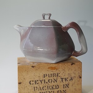 Puede incluir: Una tetera de cerámica con un diseño geométrico en tonos grises y morados. La tetera está sentada sobre una caja de madera con el texto "PURE CEYLON TEA PACKED IN CEYLON".