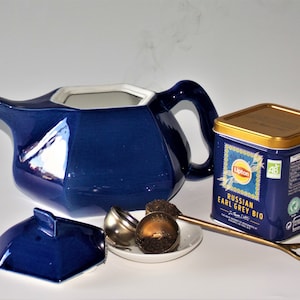 Peut inclure: Une théière en céramique bleue avec un couvercle et une tasse assortie. La théière a une forme hexagonale et un glaçage brillant. Un infuseur à thé en argent est sur une soucoupe blanche avec des feuilles de thé en vrac. Une boîte de thé Lipton Russian Earl Grey Bio est sur la table.