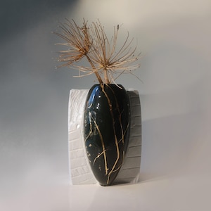 Peut inclure: Un vase en céramique vert foncé avec des accents dorés et un glaçage craquelé. Le vase est posé sur une base en céramique blanche et contient des fleurs séchées.