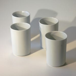 Op de afbeelding: Vier witte keramische bekers zijn gerangschikt op een wit oppervlak. De cilindrische bekers hebben een gladde, glanzende afwerking en een eenvoudig, minimalistisch ontwerp. De verlichting creëert zachte schaduwen.