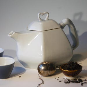 May include: White ceramic teapot with a geometric design and a heart-shaped lid. The teapot is sitting on a white surface with a tea strainer and loose leaf tea.