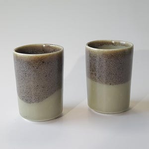 May include: Two ceramic mugs with a speckled brown and green glaze. The mugs are cylindrical in shape and have a rounded bottom.