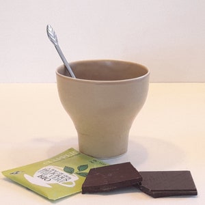 Peut inclure: Un bol en céramique beige avec une cuillère en argent en forme de feuille posée sur le bord. Le bol est posé sur une surface blanche avec un sachet de thé vert et deux morceaux de chocolat noir.