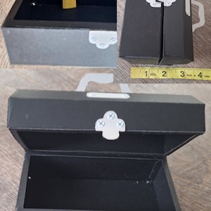 K&ouml;nnte beinhalten: Eine kleine schwarze Schachtel aus Pappe mit einem silbernen Verschluss und einem Griff. Die Schachtel ist ge&ouml;ffnet und misst etwa 10 cm in der L&auml;nge.