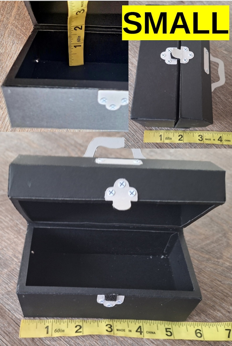 K&ouml;nnte beinhalten: Eine kleine schwarze Schachtel aus Pappe mit einem silbernen Verschluss und einem Griff. Die Schachtel ist ge&ouml;ffnet und misst etwa 10 cm in der L&auml;nge.