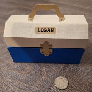 K&ouml;nnte beinhalten: Eine kleine, wei&szlig;e und blaue Holzkiste mit goldenem Griff und Verschluss. Die Kiste hat ein Namensschild auf der Vorderseite mit der Aufschrift "LOGAN".