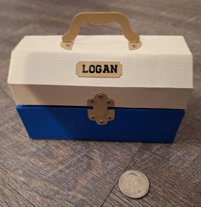 K&ouml;nnte beinhalten: Eine kleine, wei&szlig;e und blaue Holzkiste mit goldenem Griff und Verschluss. Die Kiste hat ein Namensschild auf der Vorderseite mit der Aufschrift "LOGAN".