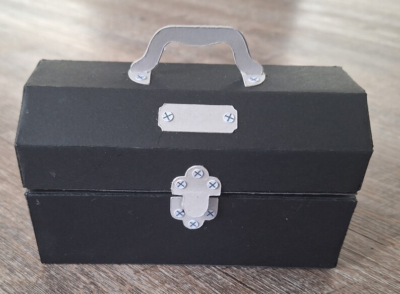 K&ouml;nnte beinhalten: Eine schwarze Pappwerkzeugbox mit einem silbernen Griff und einem silbernen Verschluss. Die Werkzeugbox hat ein kleines silbernes Etikett auf der Vorderseite.