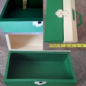 K&ouml;nnte beinhalten: Eine gr&uuml;ne und cremefarbene Schachtel aus Pappe mit einem Scharnierdeckel und einem Verschluss. Die Schachtel ist mit "MEDIUM" beschriftet. Die Schachtel ist 20 cm lang und 10 cm breit.
