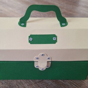 K&ouml;nnte beinhalten: Eine gr&uuml;ne und cremefarbene Pappwerkzeugbox mit Griff und Riegel. Die Werkzeugbox hat ein leeres Etikett auf der Vorderseite.
