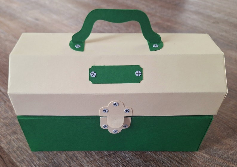 K&ouml;nnte beinhalten: Eine gr&uuml;ne und cremefarbene Pappwerkzeugbox mit Griff und Riegel. Die Werkzeugbox hat ein leeres Etikett auf der Vorderseite.