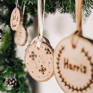 May include: Wooden Christmas decorations with a variety of designs, including snowflakes, a wreath, and the word "Harris". The decorations are hanging from white ribbon on a green pine branch.
