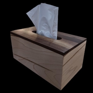 May include: A rectangular wooden tissue box with a light-colored body and a dark brown top border. White tissues are partially pulled from a circular opening. The box is made of wood and has a natural finish.