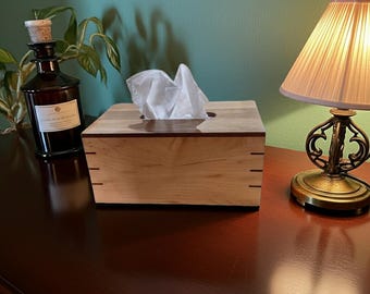 May include: A wooden tissue box with a light wood grain finish and a dark wood trim. The box is square with a rectangular opening for tissues. White tissues are visible. A brown bottle with a cork stopper and a lamp are also in the image.