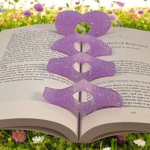 May include: Four lavender-colored book page holders with a glittery finish are arranged on an open book. The book is surrounded by a field of colorful flowers. The page holders are heart and wing shaped.