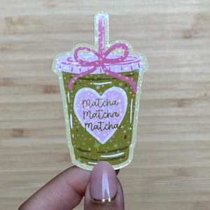 Może przedstawiać: Holograficzna naklejka z napojem matcha. Kubek jest zielony z różową pokrywką i słomką. Różowe serce ze słowami "Matcha Matcha Matcha" znajduje się na środku. Naklejka ma błyszczący efekt.