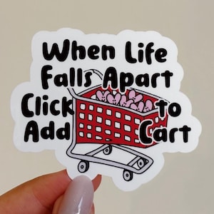 May include: A white sticker with black text that reads "When life falls apart click add to cart". A red shopping cart with pink hearts inside is illustrated next to the text.