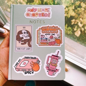 May include: A collection of colorful stickers featuring illustrations of books, coffee, and a cat holding a skull. The stickers have text that reads "One More Chapter!" "Notes" "Cozy Bookish Weekend" "Admit One" "The Cat Lady" "Fairytales" "I Like My Spicy" "Finish Your TBR Fuel" "Iced Coffee"