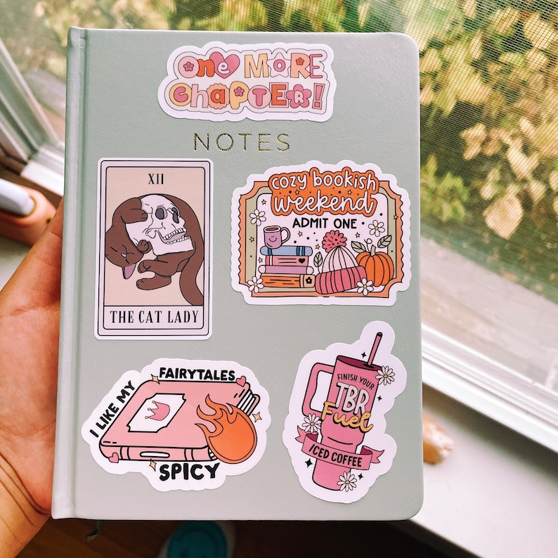 May include: A collection of colorful stickers featuring illustrations of books, coffee, and a cat holding a skull. The stickers have text that reads "One More Chapter!" "Notes" "Cozy Bookish Weekend" "Admit One" "The Cat Lady" "Fairytales" "I Like My Spicy" "Finish Your TBR Fuel" "Iced Coffee"