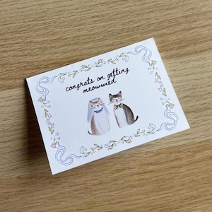 Pu&ograve; includere: Un biglietto d'auguri rettangolare bianco con un'illustrazione ad acquerello di due gatti. Un gatto indossa un velo e una collana blu, l'altro un papillon verde. Il testo "congrats on getting meowwied" &egrave; circondato da un bordo a nastro blu.