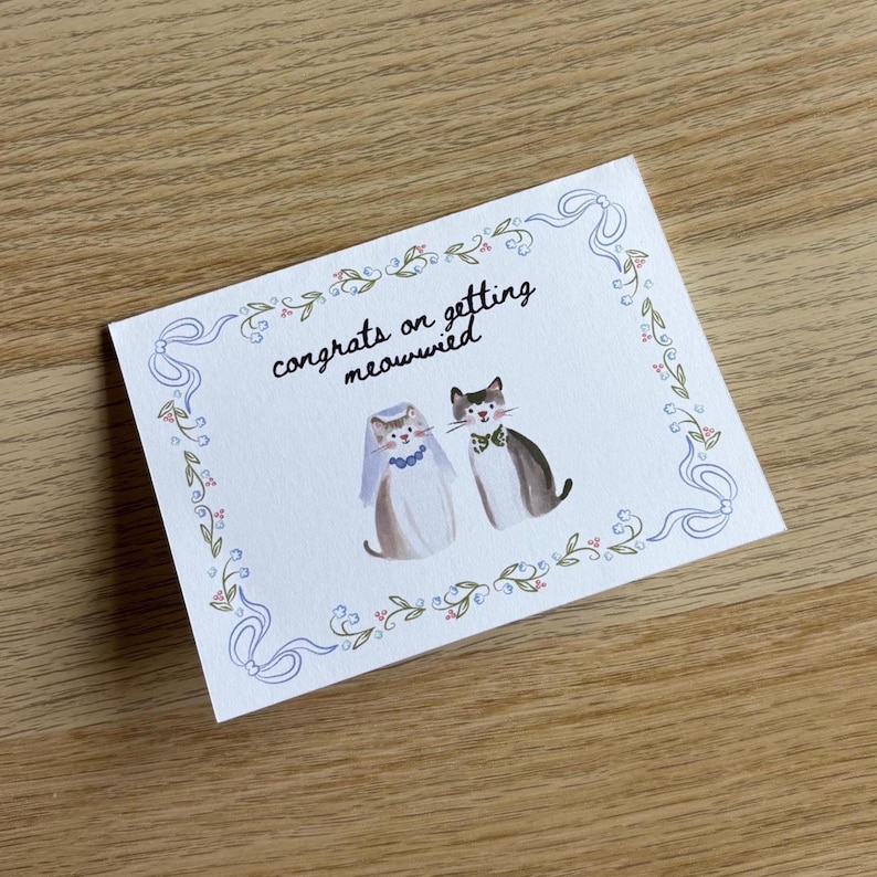 Pu&ograve; includere: Un biglietto d'auguri rettangolare bianco con un'illustrazione ad acquerello di due gatti. Un gatto indossa un velo e una collana blu, l'altro un papillon verde. Il testo "congrats on getting meowwied" &egrave; circondato da un bordo a nastro blu.