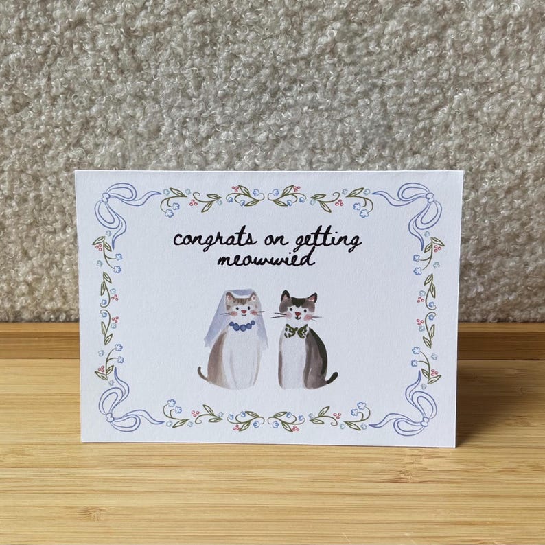 Pu&ograve; includere: Un biglietto d'auguri rettangolare bianco con un bordo decorativo di nastri blu e piccoli fiori. Il testo "congrats on getting meowwied" si trova sopra un'illustrazione di due gatti, uno che indossa un velo blu e l'altro un papillon verde.