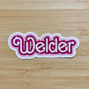May include: A sticker with the word "Welder" in white, outlined in pink and magenta, with a holographic sparkle effect. The sticker is on a light brown wooden surface.