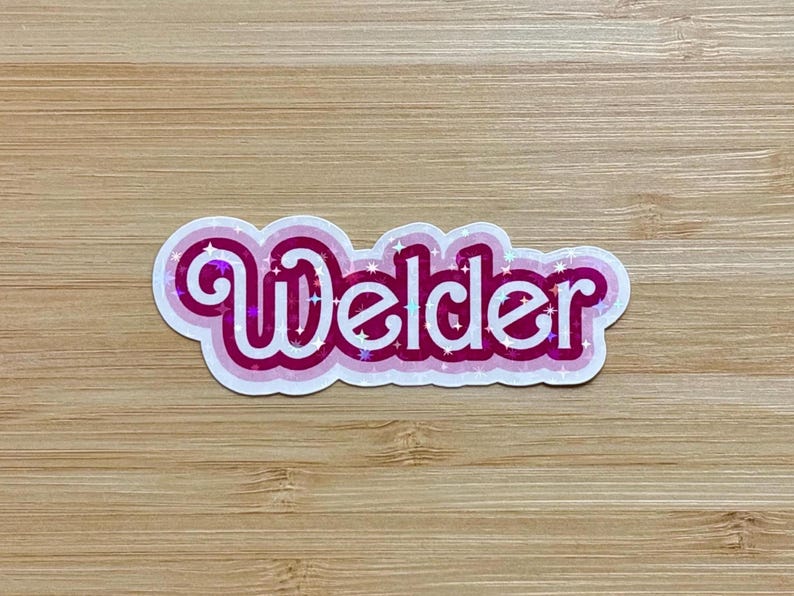 May include: A sticker with the word "Welder" in white, outlined in pink and magenta, with a holographic sparkle effect. The sticker is on a light brown wooden surface.