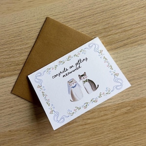Pu&ograve; includere: Un biglietto d'auguri bianco con busta marrone che presenta un'illustrazione ad acquerello di due gatti, uno con velo e l'altro con papillon. Il testo recita congrats on getting meowwied, circondato da un bordo di nastri blu e fiori.