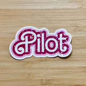 May include: A pink and white sticker with the word "Pilot" in a retro font. The sticker has a holographic, glittery effect and is outlined in white. The background is a light brown wooden surface.