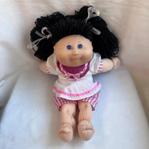 May include: A doll with a soft fabric body, light skin tone, and bright blue eyes. It has thick black yarn hair tied with white ribbons. The doll wears a white top with pink striped sleeves and a purple collar, paired with matching pink striped shorts.