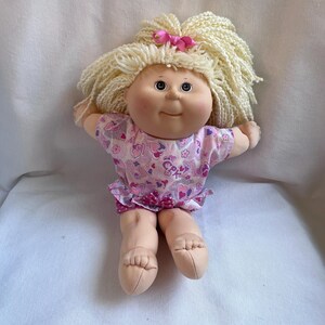 Può includere: Una bambola dal corpo morbido con pelle chiara, guance rotonde e fossette. La bambola ha capelli in lana bionda con un piccolo fiocco rosa. Indossa una maglietta rosa a maniche corte con un motivo di cuori, fiori e le lettere CPK.