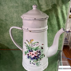 Puede incluir: Una cafetera alta de esmalte gris claro con tapa a juego, asa curva y pico largo. El cuerpo presenta un diseño floral pintado a mano con flores moradas, rosas y amarillas rodeadas de hojas verdes, con un ligero desgaste en los bordes.
