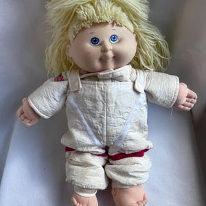 May include: A doll with pale skin, blue eyes, and light yellow yarn hair. The doll wears a white, textured, long-sleeved jumpsuit with a small American flag patch on the right shoulder and red accents around the waist and leg areas.