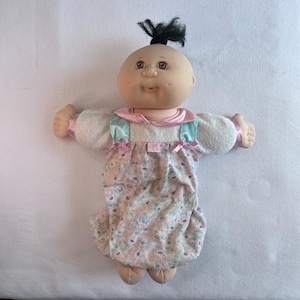 Könnte beinhalten: Eine Puppe mit rundem, hellhäutigem Gesicht, braunen Augen und einer einzelnen schwarzen Haarsträhne, die oben zusammengebunden ist. Die Puppe trägt ein weißes Langarmshirt mit rosa Satinsaum und einen rosa Strampler mit Blumenmuster und einem kleinen Etikett mit der Aufschrift opk.