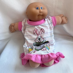 Può includere: Una bambola calva con occhi azzurri e carnagione abbronzata indossa un vestito bianco a maniche corte con sottili righe orizzontali blu e rosa. L'abito presenta una grafica di un coniglio rosa e un orlo a balze rosa con polsini abbinati.