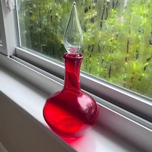 May include: A vibrant red glass decanter with a rounded base and a slender neck, topped with a clear, teardrop-shaped glass stopper. The vessel features a smooth, polished surface that catches the light, creating a translucent effect.