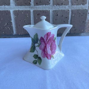 May include: A white porcelain creamer with a lid, featuring a large pink rose and green leaves. The creamer has a square base and a curved handle. The creamer is approximately 4 inches tall.
