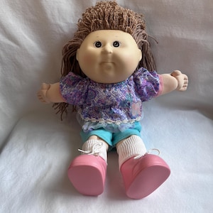 Puede incluir: Una muñeca de piel clara, cabello de lana marrón y ojos oscuros. La muñeca viste una blusa morada de manga corta con un borde floral blanco, pantalones cortos de color verde azulado, calcetines blancos acanalados y zapatos de color rosa.